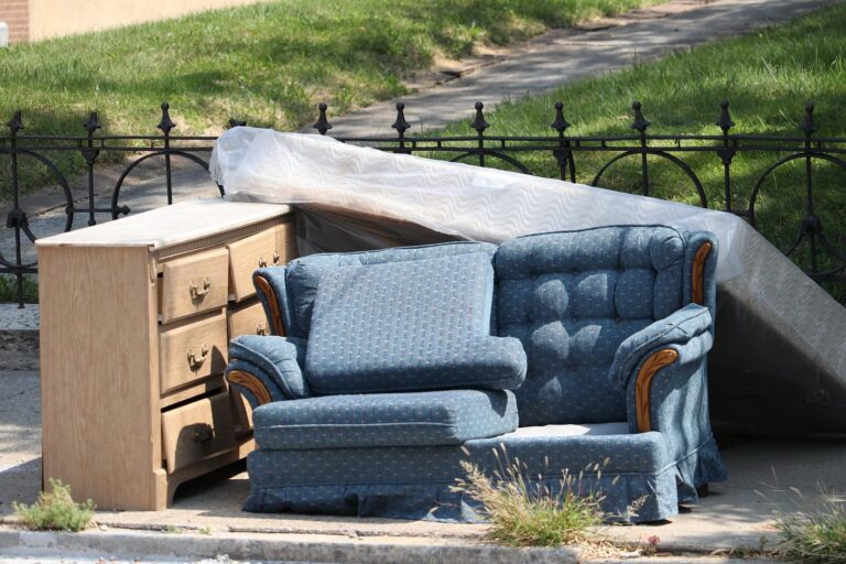 Old mattress in plastic leaning on a fence behind a blue sofa and dresser left on the kerb for collection