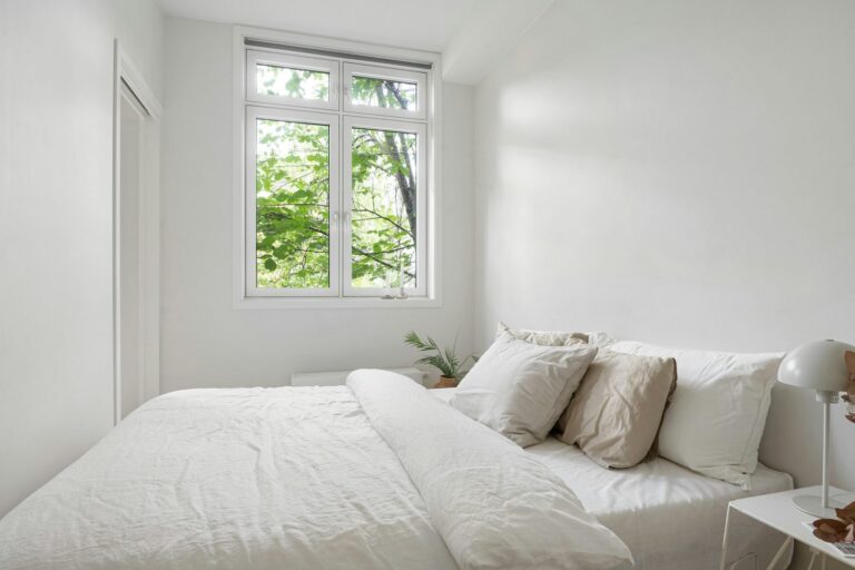 Airy white bedroom with linen bedding and a window looking out to green summer leaves
